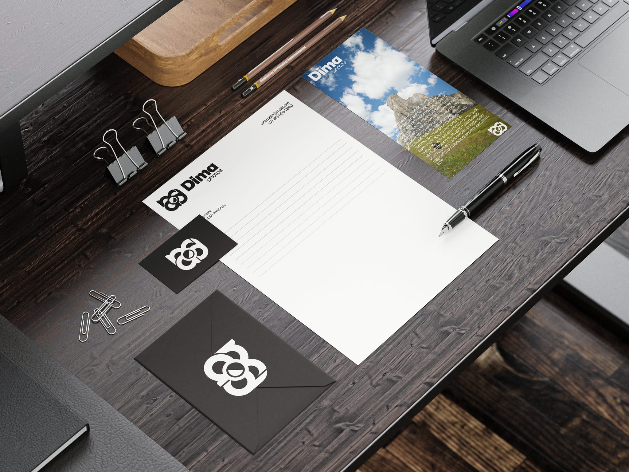 La cancelleria di Dima photos su una scrivania di legno scuro: la carta intestata col logo, la busta nera col monogramma bianco, il biglietto e un volantino con la foto di una montagna.