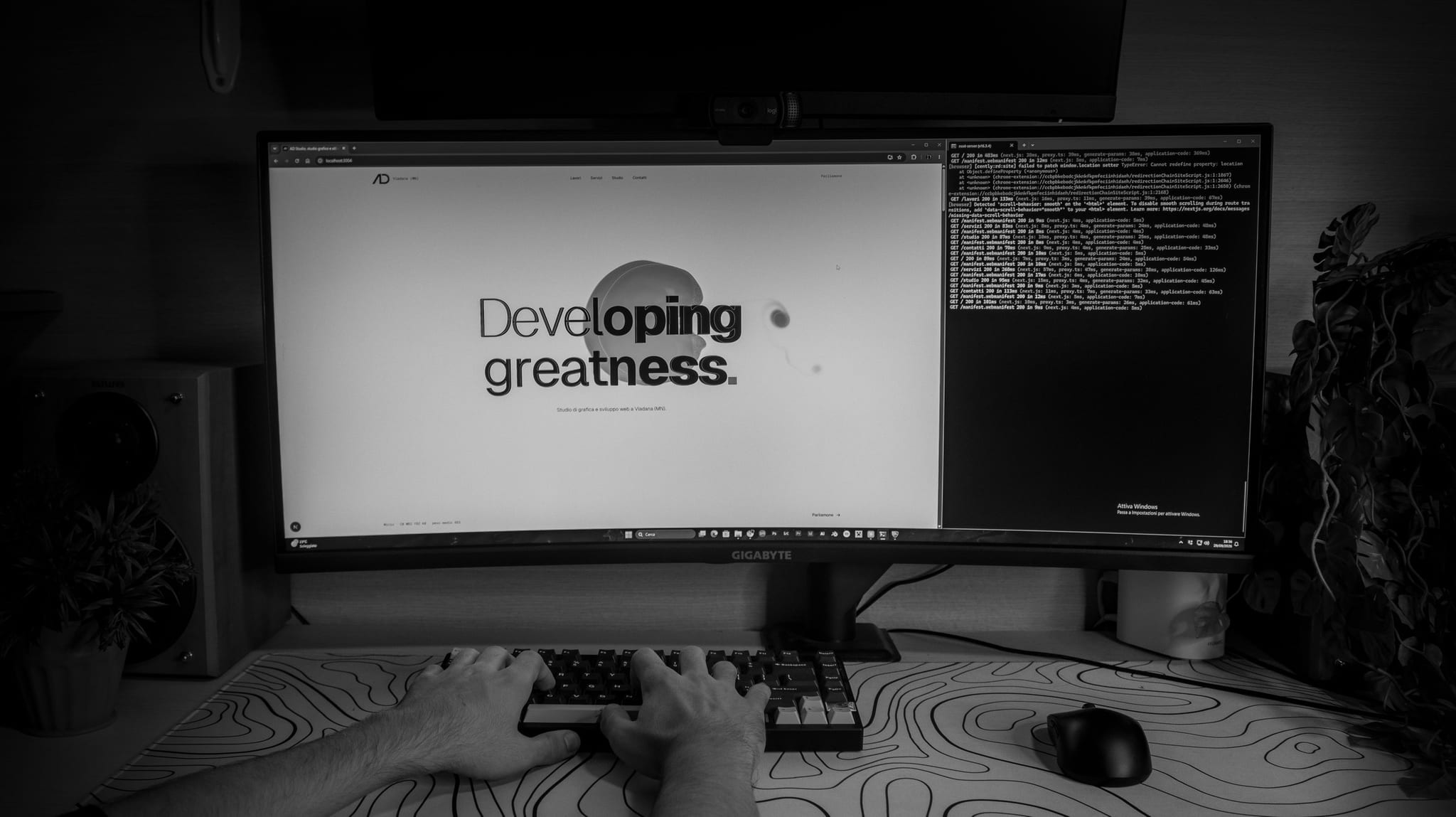 La nostra scrivania: sul monitor curvo c’è l’apertura di questo sito, «Developing greatness.», con accanto la finestra del terminale; in primo piano le mani sulla tastiera, su un tappetino con le curve di livello.