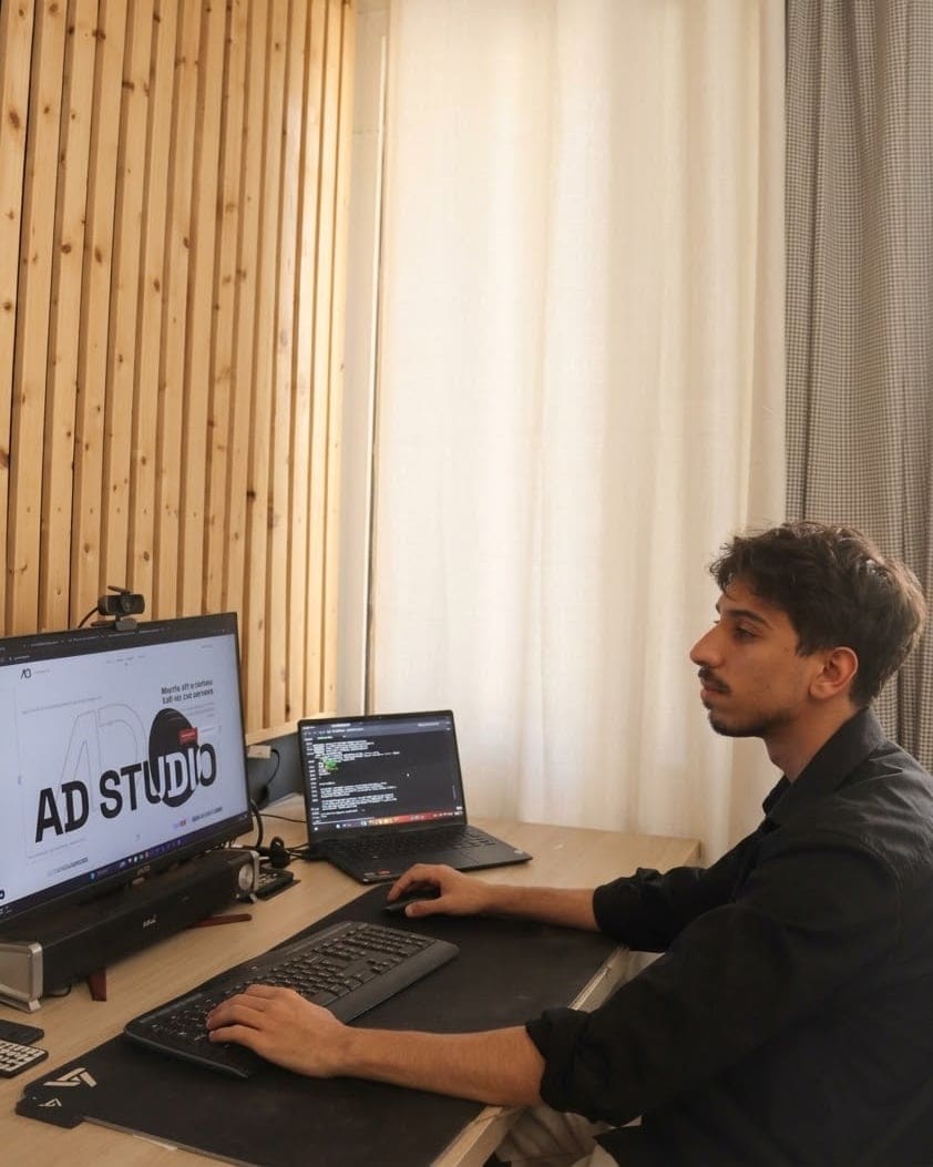 Dennis en su escritorio, de perfil, una mano en el teclado y otra en el ratón. En la pantalla grande está la página de apertura de este sitio; en el portátil de al lado, unas líneas de código.